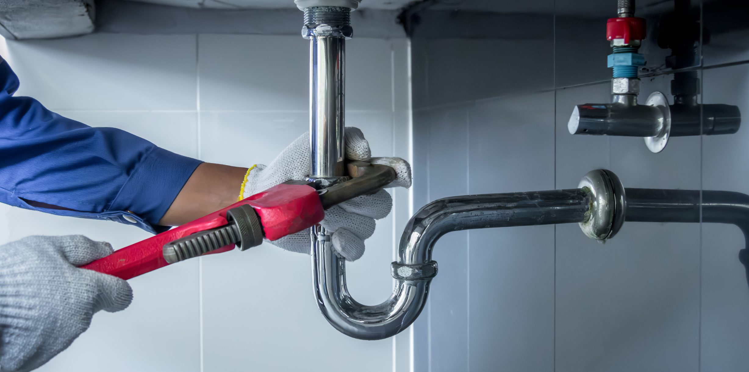 Plumber tightening a chrome drain pipe with a bright red wrench under a sink
