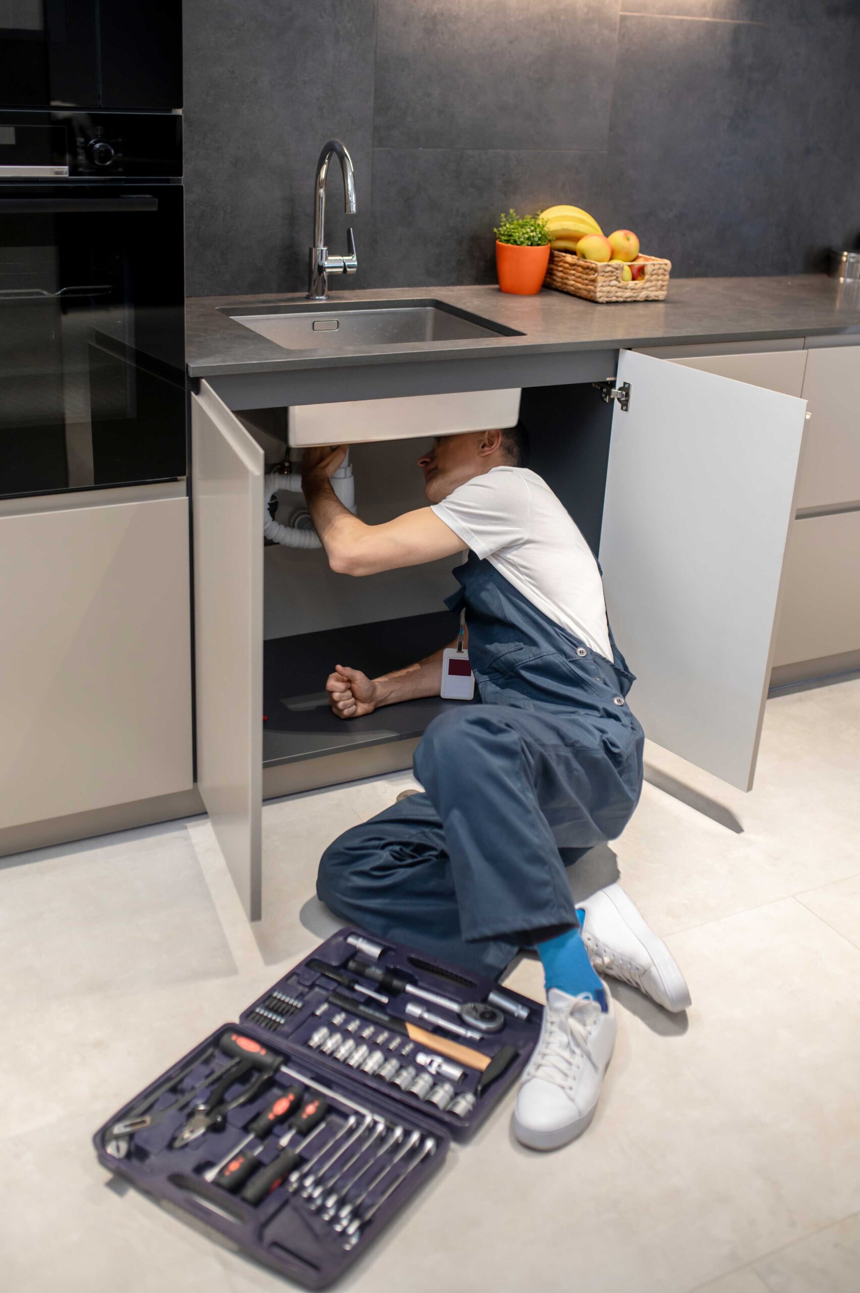 Plumber working under a modern kitchen sink with an organized tool case open beside him
